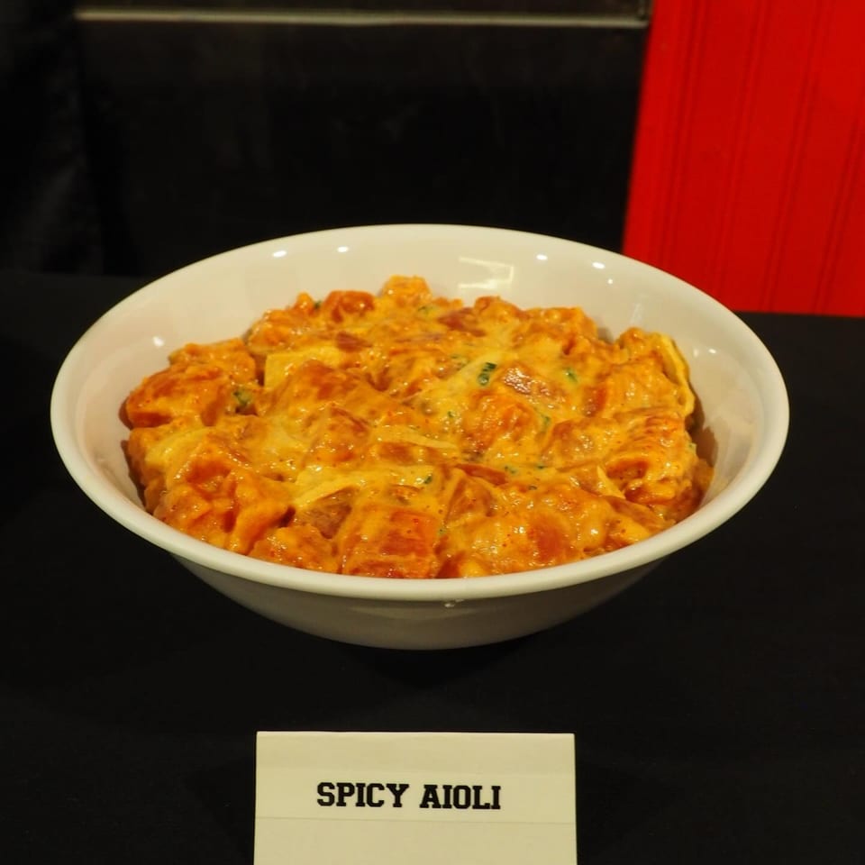 Bowl of golden fried bites coated in spicy aioli, displayed on a white plate against a dark background.